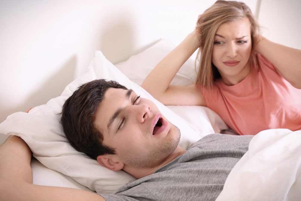 Casal deitado na cama, homem roncando enquanto dorme e mulher acordado ao seu lado